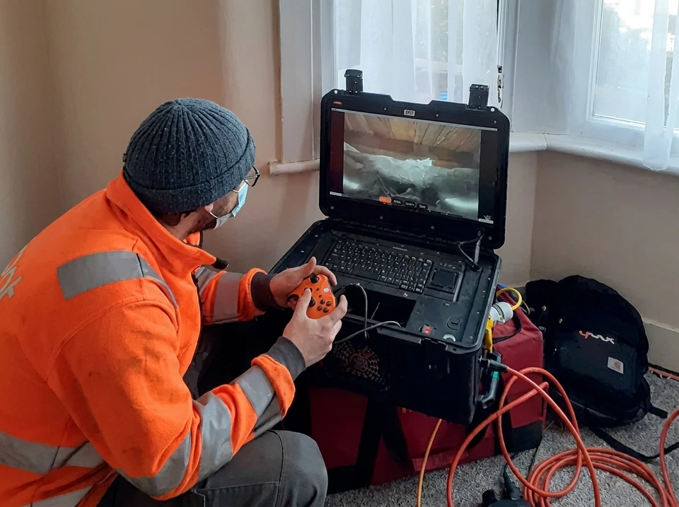 Technicien en veste orange haute visibilité et masque chirurgical, opérant une caméra d'inspection robotisée via une manette de jeu Xbox dans un espace intérieur