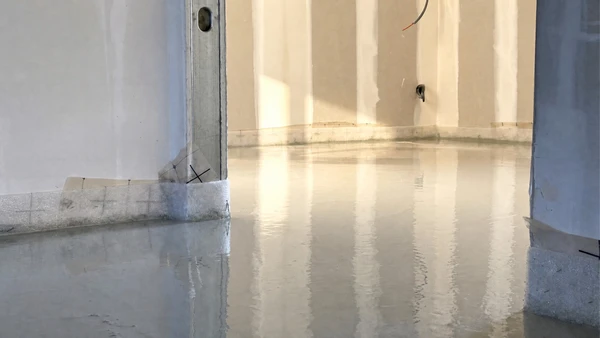 Ouvrier en bottes de caoutchouc appliquant une chape fluide autonivelante grise fraîchement coulée à l'aide d'une règle de maçon métallique guidée par des rails de nivellement en aluminium. Surface de béton lisse et réfléchissante caractéristique d'une chape liquide anhydrite ou ciment. Intérieur de chantier en construction avec murs en plaques de plâtre partiellement enduits, plots de nivellement jaunes visibles, aspect professionnel de ragréage intérieur.