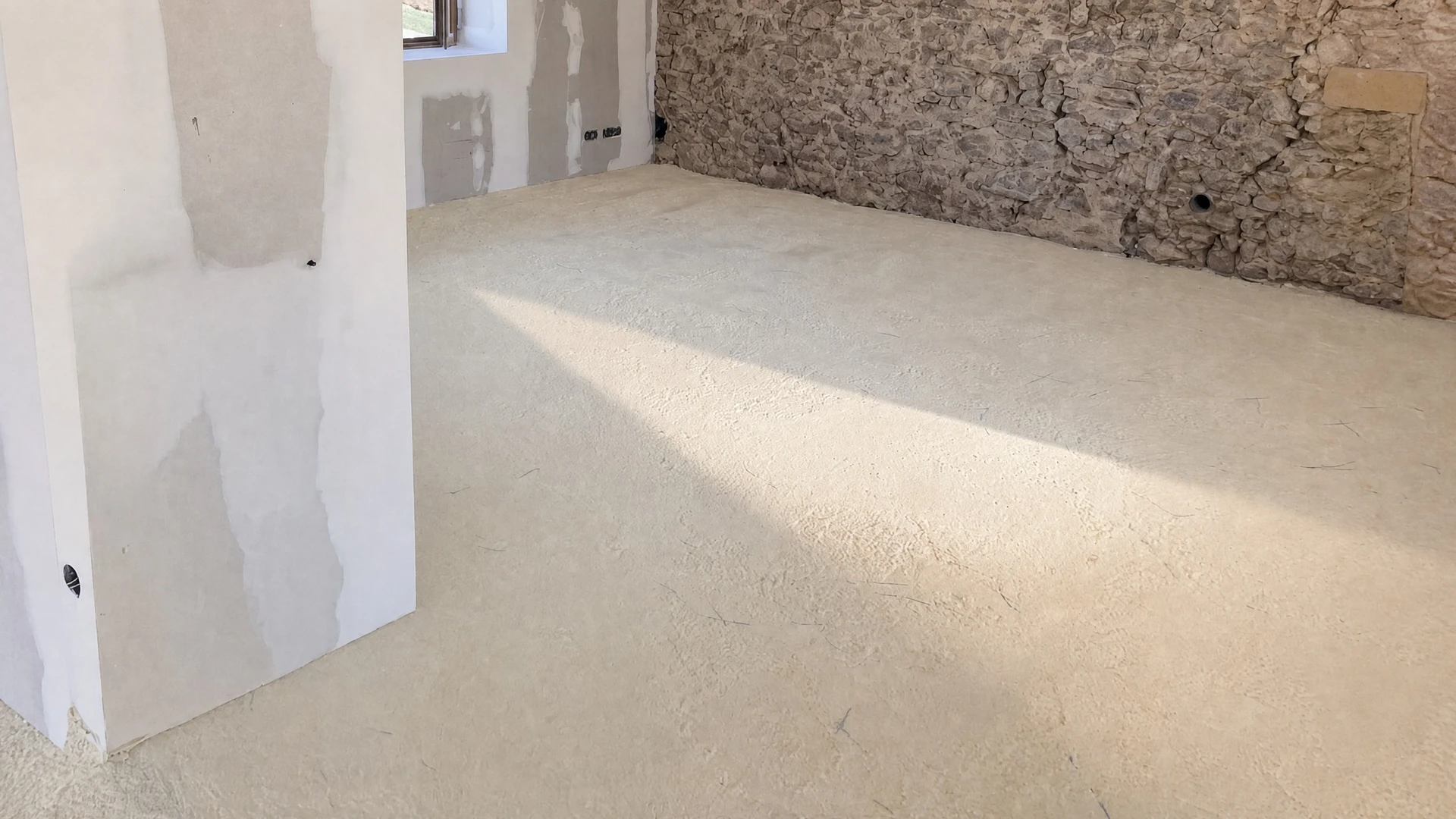 Ouvrier en tenue de chantier standard appliquant une chape fluide ciment autonivelante à l'aide d'une règle de maçon sur un plancher chauffant en construction. Le sol montre une surface lisse, gris clair, fraîchement coulée et brillante. Plots de nivellement jaunes visibles en arrière-plan. Murs en plaques de plâtre partiellement enduits. Tubes de chauffage au sol partiellement visibles. La scène capture le geste professionnel du ragréage/lissage, incarnant le savoir-faire d'Euro Chape Fluide dans la pose de chapes fluides précises et régulières.